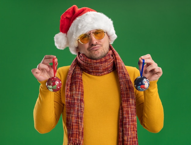 Jeune homme à col roulé jaune et bonnet de Noel portant des lunettes tenant des jouets pour arbre de Noël à la confusion et mécontent debout sur mur vert