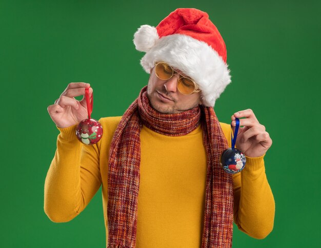 Jeune homme en col roulé jaune et bonnet de Noel portant des lunettes tenant des jouets pour arbre de Noël en les regardant avec un visage sérieux debout sur un mur vert