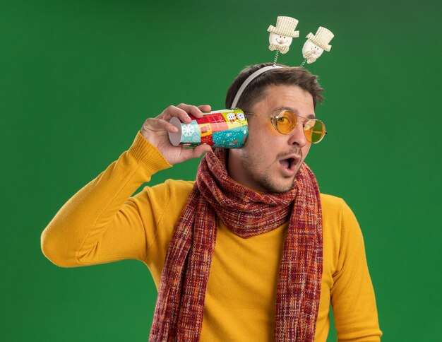 Jeune homme à col roulé jaune avec écharpe chaude et lunettes portant une jante drôle sur la tête tenant une tasse colorée sur l'oreille en essayant d'écouter les commérages debout sur le mur vert