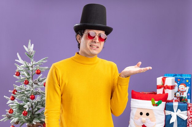 Jeune homme en col roulé jaune et lunettes portant un chapeau noir avec le bras d'être confus debout à côté d'un arbre de Noël et présente sur mur violet