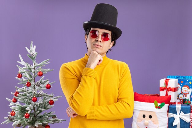 Jeune homme à col roulé jaune et lunettes portant un chapeau noir à côté avec la main sur le menton pensant debout à côté d'un arbre de Noël et présente sur fond violet