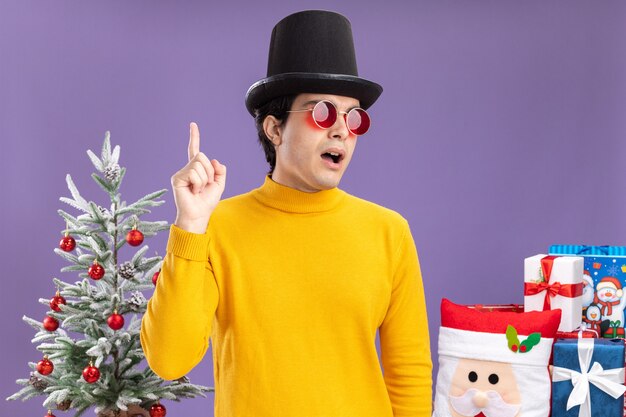 Jeune homme à col roulé jaune et lunettes portant un chapeau noir à côté surpris montrant l'index ayant une excellente idée debout à côté d'un arbre de Noël et présente sur mur violet