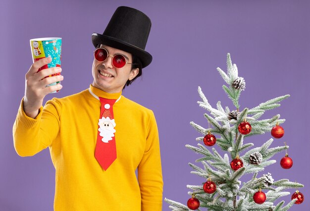 Jeune homme à col roulé jaune et lunettes portant un chapeau noir et une cravate drôle tenant une tasse de papier coloré loking à elle avec le sourire sur le visage debout à côté d'un arbre de Noël sur un mur violet