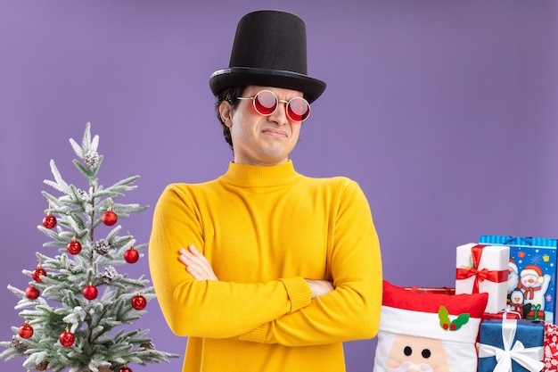 Jeune homme à col roulé jaune et lunettes portant un chapeau noir regardant la caméra mécontent avec les bras croisés sur la poitrine debout à côté d'un arbre de Noël et présente sur fond violet