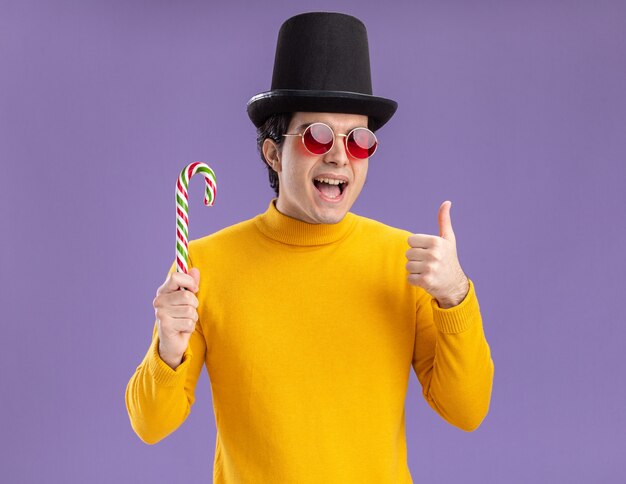 Jeune homme à col roulé jaune et lunettes portant un chapeau noir tenant la canne en bonbon souriant joyeusement montrant les pouces vers le haut debout sur le mur violet