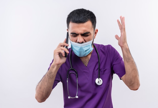 Jeune homme en colère médecin portant des vêtements de chirurgien violet et masque médical stéthoscope speakes sur téléphone levant la main sur un mur blanc isolé