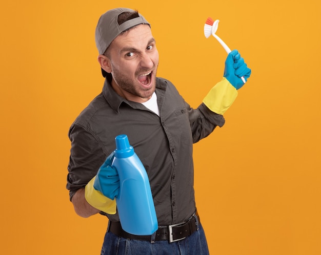 Jeune homme en colère de nettoyage portant des vêtements décontractés et une casquette de gants en caoutchouc tenant une brosse de nettoyage et une bouteille avec des produits de nettoyage criant avec une expression agressive debout sur un mur orange