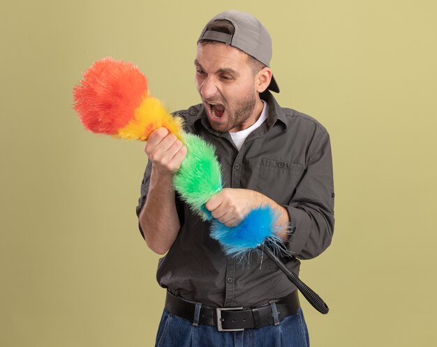 Jeune homme en colère de nettoyage portant des vêtements décontractés et une casquette tenant un plumeau coloré en criant avec une expression agressive debout sur un mur vert