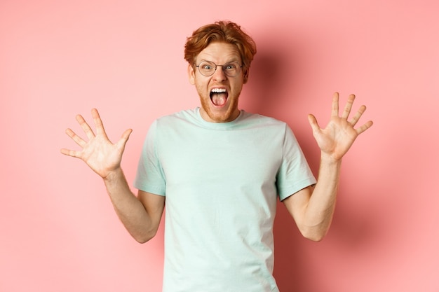 Jeune homme en colère et sous pression, perdre son sang-froid, écarter les mains sur le côté et crier avec un visage furieux, debout dans des lunettes et un t-shirt sur fond rose.