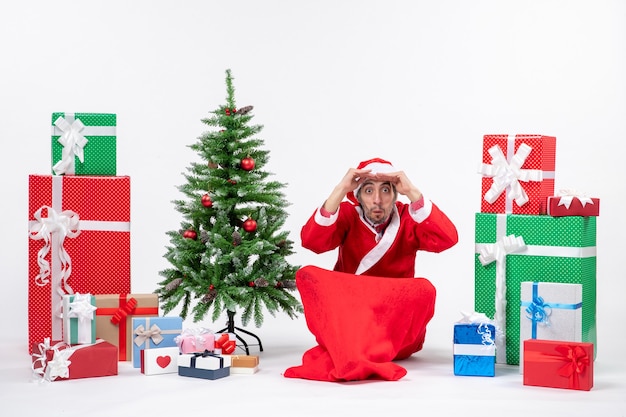 Jeune homme concentré célébrer le nouvel an ou les vacances de Noël assis sur le sol près de cadeaux et arbre de Noël décoré