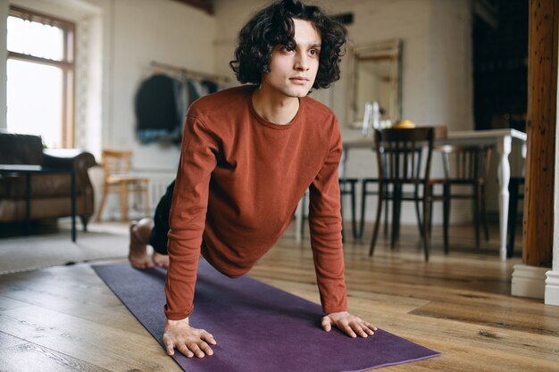 Jeune homme confiant autodéterminé aux cheveux bouclés faisant planche sur un tapis de fitness pendant l'entraînement du matin à la maison en raison de la distance sociale.