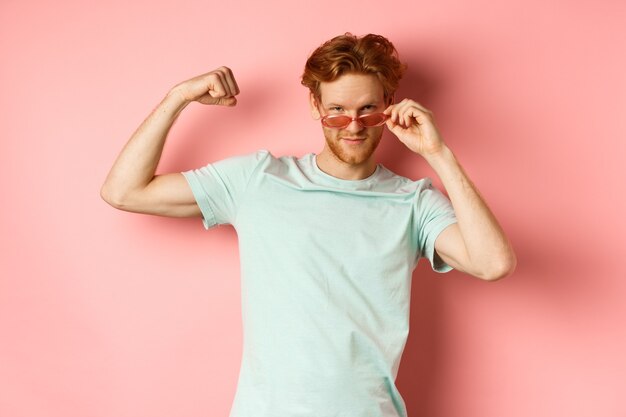 Jeune homme confiant aux cheveux rouges portant des lunettes de soleil d'été et un t-shirt montrant un corps fort et en forme...