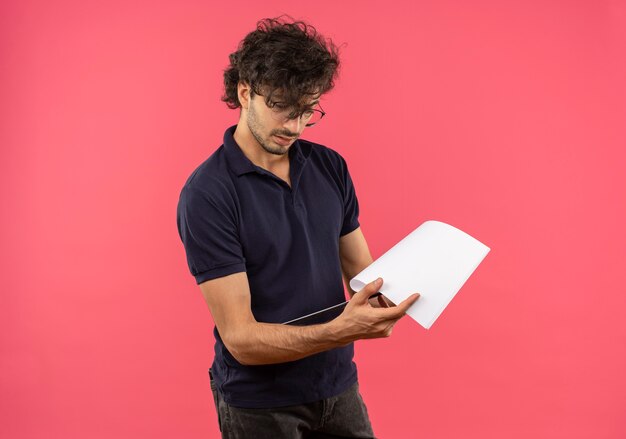 Jeune homme confiant en chemise noire avec des lunettes optiques détient et regarde le presse-papiers isolé sur mur rose