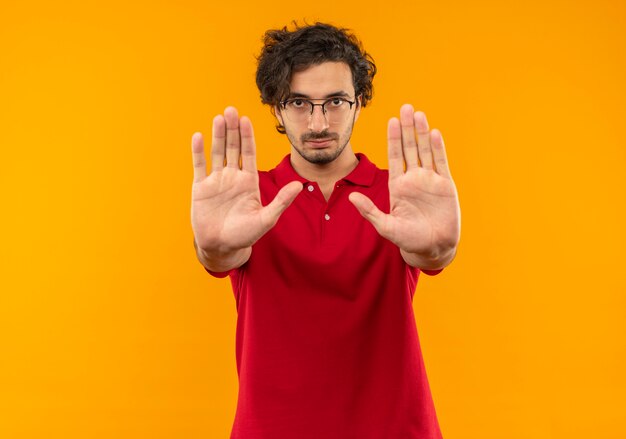 Jeune homme confiant en chemise rouge avec des gestes de lunettes optiques s'arrêtent avec les mains isolées sur le mur orange