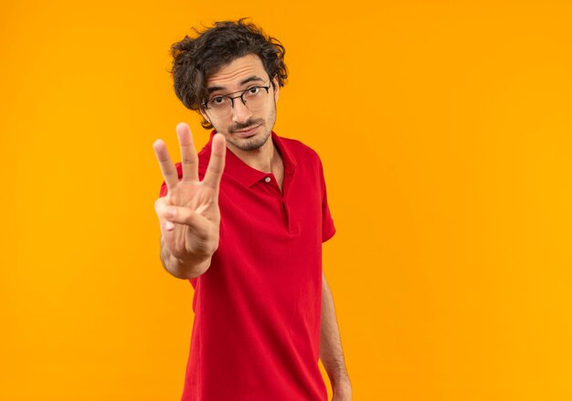 Jeune homme confiant en chemise rouge avec des gestes de lunettes optiques trois signe de la main isolé sur un mur orange