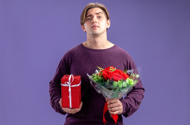 Jeune homme confiant le jour de la Saint-Valentin tenant une boîte-cadeau avec bouquet isolé sur fond bleu