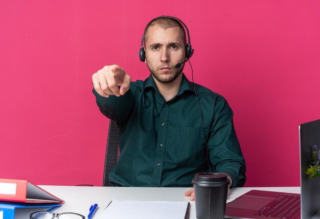 Jeune homme confiant opérateur de centre d'appels portant un casque assis au dask avec des outils de bureau vous montrant un geste