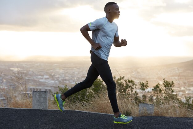 Un jeune homme confiant porte un t-shirt blanc, des leggings noirs et des baskets, court seul sur l'autoroute, respire profondément, démontre son endurance, aime le matin. Gens, course, concept de mode de vie