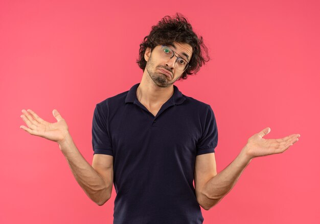 Jeune homme confus en chemise noire avec des lunettes optiques tient les mains et semble isolé sur un mur rose