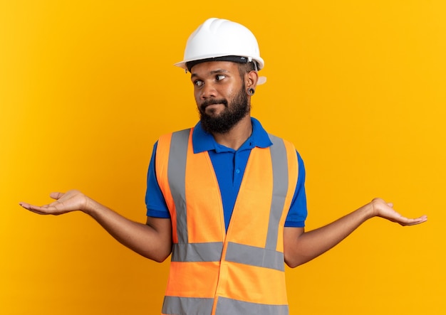 Jeune homme de constructeur afro-américain confus en uniforme avec un casque de sécurité tenant ses mains ouvertes regardant le côté isolé sur fond orange avec espace de copie