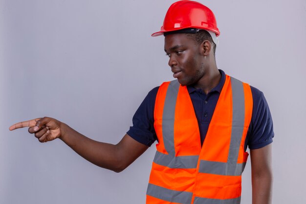 Jeune homme de constructeur afro-américain portant un gilet de construction et un casque de sécurité à côté et pointant avec le doigt sur le côté debout