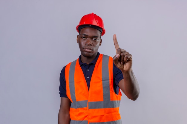 Jeune homme constructeur afro-américain portant un gilet de construction et un casque de sécurité debout avec un doigt d'avertissement de danger sur blanc isolé