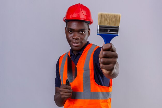 Jeune homme de constructeur afro-américain portant un gilet de construction et un casque de sécurité montrant un pinceau qui s'étend à la confiance sur blanc isolé