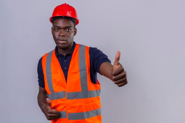 Jeune homme de constructeur afro-américain portant un gilet de construction et un casque de sécurité montrant le pouce debout