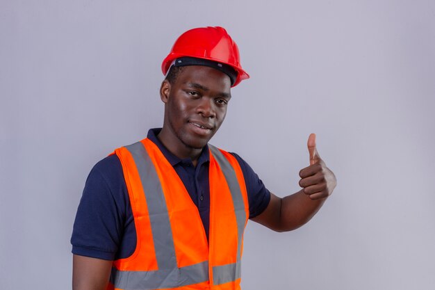 Jeune homme de constructeur afro-américain portant un gilet de construction et un casque de sécurité montrant les pouces vers le haut avec le sourire sur le visage debout