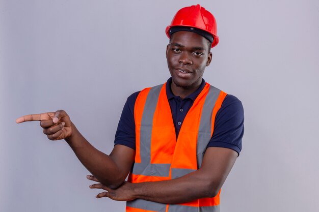Jeune homme constructeur afro-américain portant un gilet de construction et un casque de sécurité pointant avec le doigt sur le côté à la confiance sur blanc isolé