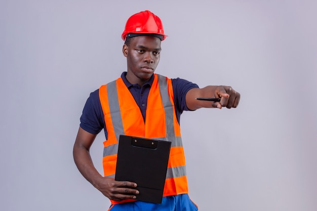 Jeune homme de constructeur afro-américain portant un gilet de construction et un casque de sécurité pointant l'index à distance avec une expression sérieuse debout