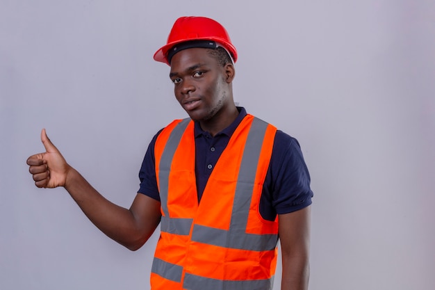 Jeune homme de constructeur afro-américain portant un gilet de construction et un casque de sécurité avec le sourire sur le visage montrant le pouce debout