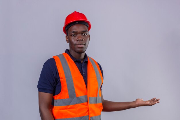 Jeune homme de constructeur afro-américain portant un gilet de construction et un casque de sécurité avec un visage sérieux présentant avec la main et la paume sur blanc isolé