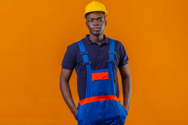 Photo gratuite jeune homme de constructeur afro-américain portant un uniforme de construction et un casque de sécurité debout avec ses mains dans les poches avec une expression sérieuse confiante sur orange