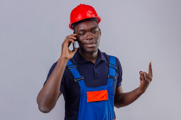 Jeune homme de constructeur afro-américain portant l'uniforme de la construction et un casque de sécurité parlant sur téléphone mobile faisant symbole rock debout souriant