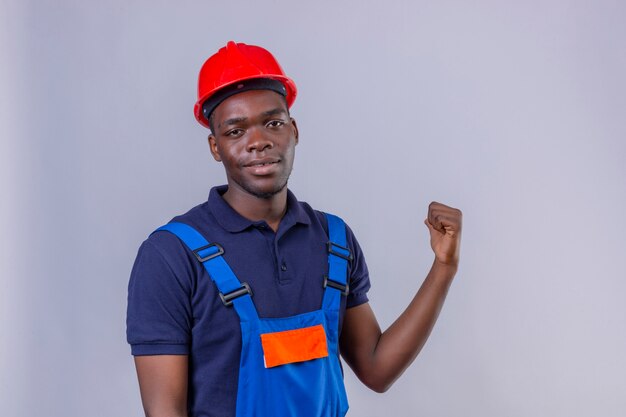 Jeune homme de constructeur afro-américain portant l'uniforme de construction et un casque de sécurité pointant vers l'arrière derrière avec la main et le pouce vers le haut souriant debout confiant