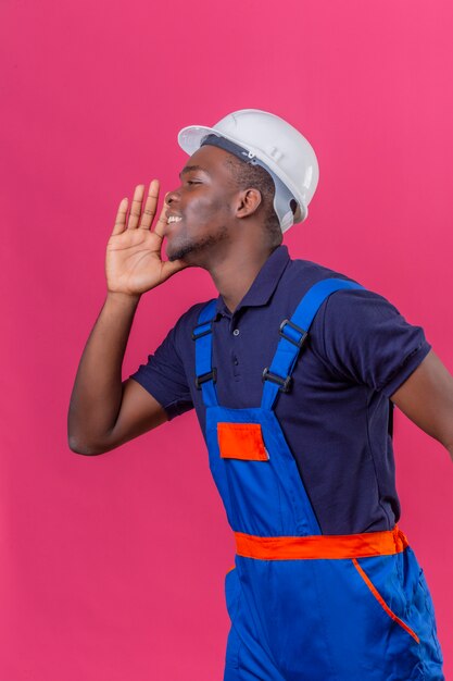 Jeune homme de constructeur afro-américain portant des uniformes de construction et un casque de sécurité criant appeler quelqu'un avec la main près de la bouche debout sur rose