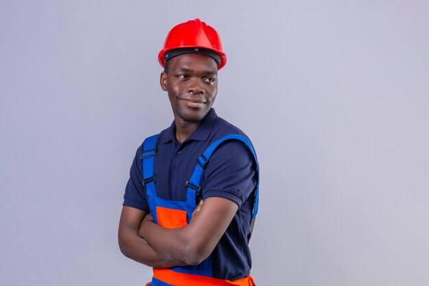 Jeune homme de constructeur afro-américain portant des uniformes de construction et un casque de sécurité debout avec les bras croisés à côté souriant debout sympathique