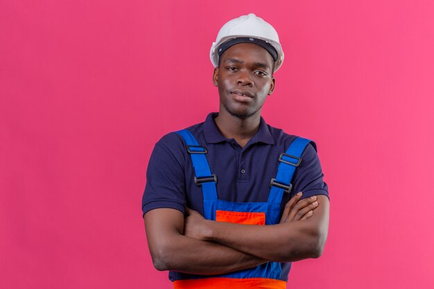 Jeune homme de constructeur afro-américain portant des uniformes de construction et un casque de sécurité debout avec les bras croisés sur la poitrine à la confiance sur rose isolé