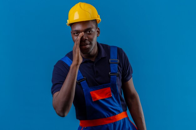 Jeune homme de constructeur afro-américain portant des uniformes de construction et un casque de sécurité debout avec une main près de la bouche en disant un secret sur bleu isolé