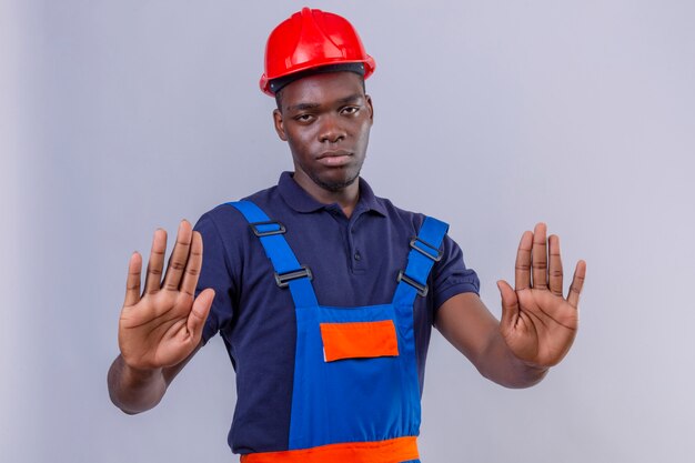 Jeune homme de constructeur afro-américain portant des uniformes de construction et un casque de sécurité debout avec les mains ouvertes faisant panneau d'arrêt avec un geste de défense d'expression sérieuse et confiante debout