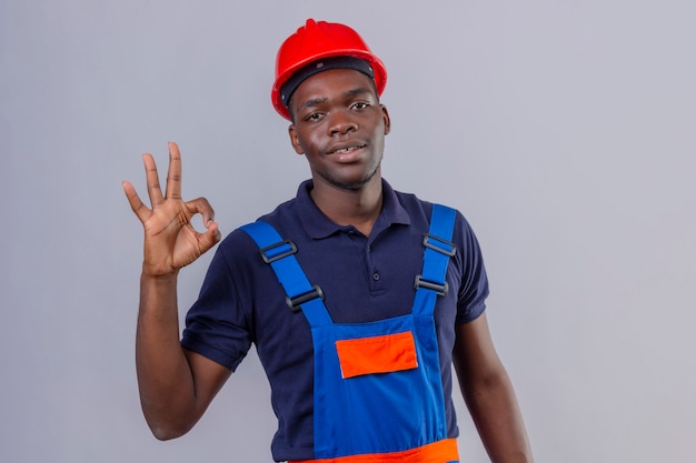 Jeune homme de constructeur afro-américain portant des uniformes de construction et un casque de sécurité avec le sourire sur le visage faisant signe ok debout
