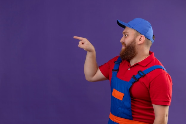 Jeune homme constructeur barbu en uniforme de construction et cap à côté en pointant avec l'index à quelque chose sur fond violet