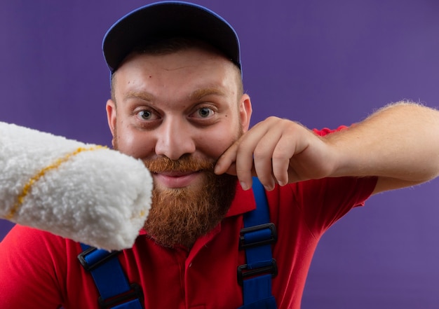 Jeune homme constructeur barbu en uniforme de construction et cap holding rouleau à peinture toucher sa moustache souriant sur fond violet