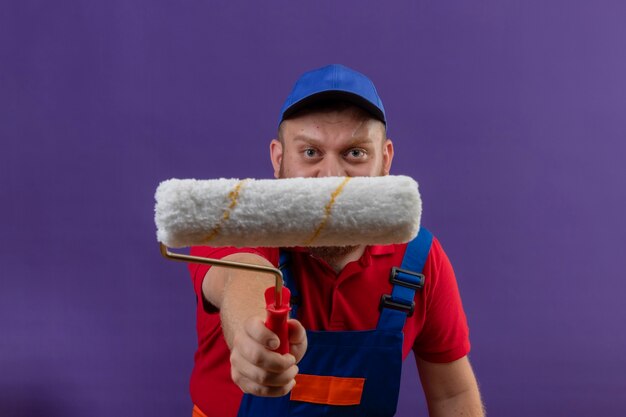 Jeune homme constructeur barbu en uniforme de construction et cap s'étendant à l'appareil photo rouleau à peinture sur fond violet