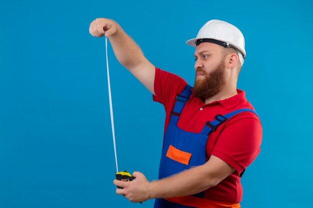 Jeune homme constructeur barbu en uniforme de construction et casque de sécurité à l'aide d'un ruban de mesure en le regardant avec un visage sérieux