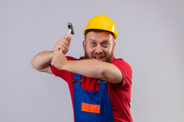 Jeune homme constructeur barbu en uniforme de construction et casque de sécurité balançant un marteau regardant la caméra en souriant