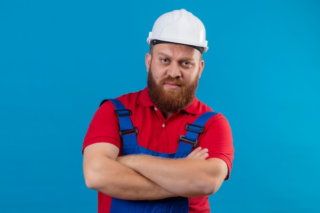 Jeune homme constructeur barbu en uniforme de construction et casque de sécurité avec les bras croisés mécontent