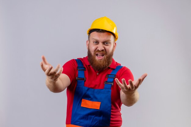 Jeune homme constructeur barbu en uniforme de construction et casque de sécurité en colère et frustré hurlant avec les bras levés