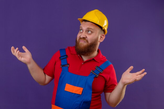 Jeune homme constructeur barbu en uniforme de construction et casque de sécurité à la confusion en haussant les épaules n'ayant pas de réponse, en écartant les bras sur fond violet